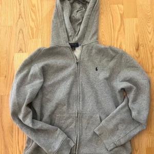 Ralph lauren grå hoodie - Klassisk grå hoodie från Polo Ralph Lauren med dragkedja och huva. Tröjan har två fickor framtill och en liten broderad logga på bröstet. Perfekt för en avslappnad stil.