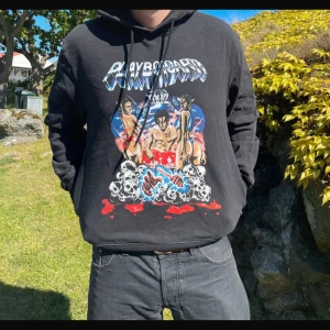Svart Playboy Carti hoodie med tryck - Säljer en svart hoodie från Playboy Carti med stort färgglatt tryck framtill och turnédatum i rött på ryggen. Tröjan har huva med snörning och är tillverkad i bomull och polyester. Perfekt för dig som gillar streetwear och musikmerch.