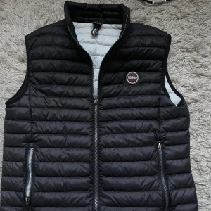 Svart dunväst från Colmar storlek 48 passar mig som har storlek M och S - Snygg svart dunväst från Colmar med quiltad design och dragkedja framtill. Västen har två fickor med dragkedja och en hög krage. Insidan är ljusgrå och har en innerficka med dragkedja. Perfekt för lager-på-lager-stil.