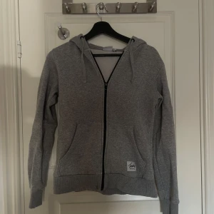 Grå hoodie med dragkedja från Andy by Frank Dandy - Säljer en klassisk grå hoodie från Andy by Frank Dandy i storlek S. Tröjan har dragkedja framtill, två fickor och huva med snören. Perfekt för en avslappnad stil och enkel att matcha med det mesta.