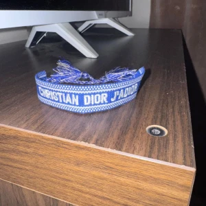 Blått armband från Christian Dior - Säljer ett blått armband från Christian Dior med texten 'J'ADIOR' broderad i vitt. Armbandet har justerbara knytband med tofsar i ändarna och är gjort i tyg med ett snyggt mönster.