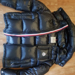Svart Moncler Montbeliard jacka - Snygg svart dunjacka från Moncler med glansig finish och klassisk logga på bröstfickan. Jackan har flera dragkedjeförsedda fickor och coola detaljer som rödvit rand längs dragkedjan. Perfekt för kalla dagar och riktigt street.