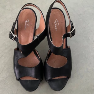 Svarta sandaler med klack från Rizzo - Säljer ett par svarta sandaler från Rizzo med öppen tå och hög klack. Skorna har breda remmar och justerbart spänne vid ankeln. Perfekta för dig som vill ha en stilren och klassisk look.