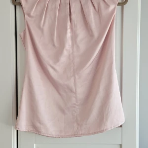 Ljusrosa satintopp med dragkedja - Säljer en ljusrosa satintopp med lätt glansig yta och diskreta veck upptill. Toppen är ärmlös och har en dold dragkedja i ryggen. Perfekt för dig som gillar en feminin och stilren look.