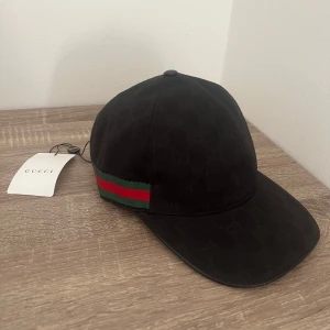 Svart keps från Gucci med GG-mönster - Snygg svart keps från Gucci med klassiskt GG-mönster i svart och röd-grön rand på sidan. Justerbar rem baktill i skinn och insida i svart tyg. Perfekt för dig som vill ha en exklusiv och trendig accessoar.