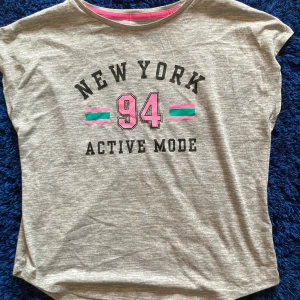 Grå sportig t-shirt H&M Active Mode - Sportig gråmelerad t-shirt från H&M med tryck 'NEW YORK 94 ACTIVE MODE' i svart och rosa, samt gröna och rosa ränder. Kortärmad modell med rund hals och rosa detalj på insidan av kragen. 