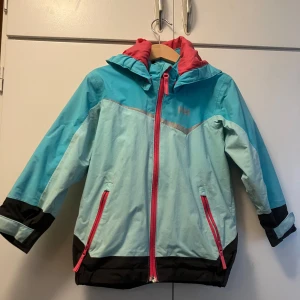 Turkos vindjacka Helly Hansen 104/4 - Snygg turkos och ljusblå vindjacka från Helly Hansen med svarta detaljer och rosa dragkedjor. Jackan har huva, långa ärmar och reflekterande HH-logga. Tillverkad i polyamid och polyester, perfekt för blåsiga dagar. Hel och ren.