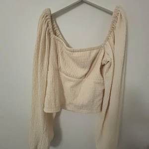 Beige croppad blus med puffärm - Superfin beige croppad blus med långa puffärmar och rynkade axlar. Toppen har en fyrkantig ringning och är gjord i ett mjukt, strukturerat tyg som känns riktigt skönt mot huden. Perfekt för dig som gillar en trendig och avslappnad stil.