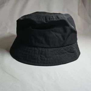 Svart bucket hat från H&M - En klassisk svart bucket hat från H&M. Hatten har rund form och bredare brätte, tillverkad i ett lätt och bekvämt material. Perfekt för att skydda mot solen och ge en avslappnad stil.