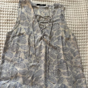 Mönstrad ärmlös blus med snörning från Bik Bok - Säljer en ljusblå och vit ärmlös topp från Bik Bok med blommigt och paisleymönster. Toppen har snörning framtill och är tillverkad i ett lätt och mjukt material. Perfekt för varma dagar!