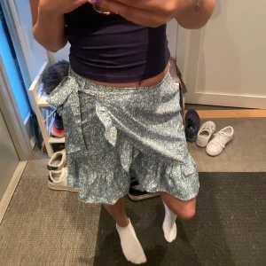 Blå blommig omlottkjol med volang - Supersöt blå kort omlottkjol med små vita blommor. Kjolen har volangkant nertill och knyts i sidan med en rosett. Perfekt för vår och sommar när du vill ha en chill och fräsch look.