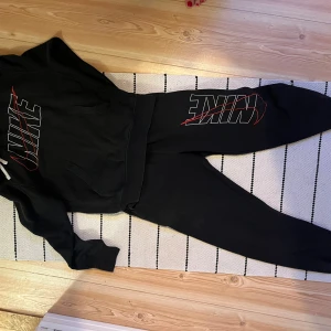 Svart Nike hoodie med stor logga XS - Svart hoodie från Nike i storlek XS med stor vit och röd logga på bröstet. Klassisk känguruficka framtill och huva med dragsko. Perfekt för chill eller träning, riktigt snygg streetstil. Mjuk och skön bomullsblandning.