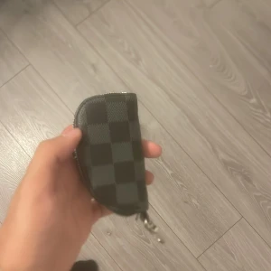 Svart-rutig väska med dragkedja - Snygg liten väska i svart med rutigt mönster och dragkedja runtom. Perfekt för nycklar, bilnycklar eller småprylar, med en metallkedja inuti för att fästa nycklar. Stilren och smidig att ha i fickan eller väskan.