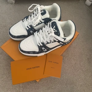 Louis Vuitton sneakers i vitt och blått - Säljer ett par exklusiva Louis Vuitton sneakers i vitt och mörkblått med tydliga LV-loggor och mönster. Skorna har snörning, rund tå och platt sula. Tillverkade i skinn och syntet, levereras med originalkartong och tillbehör.