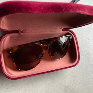 Bruna solglasögon från Gucci - Snygga solglasögon från Gucci med brunmelerad båge och mörka glas. Klassisk och tidlös design med diskret logga på sidorna. Kommer med originalfodral och tygpåse i vinröd sammet. Perfekt för sommaren 2025.
