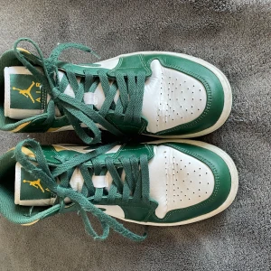 Nike Air Jordan 1 Mid grön/vit/gul - Säljer ett par Nike Air Jordan 1 Mid i grönt, vitt och gult. Skorna har klassisk snörning, vit sula och gröna detaljer med gul häl. Ikonisk Air Jordan-logga på sidan.