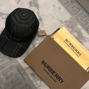 Svart keps från Burberry - Snygg svart keps från Burberry med klassiskt rutigt mönster i mörka toner. Kepsen har böjd skärm och justerbar passform. Tillverkad i en blandning av bomull och polyester. Perfekt accessoar för en stilren look.