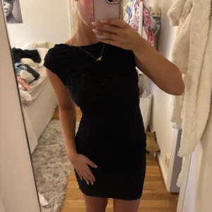 Svart kort klänning med öppen rygg - Supersnygg svart kort klänning med tight passform och öppen rygg. Klänningen har ett diskret strukturerat tyg och är ärmlös med breda axelband. Perfekt för dig som vill sticka ut med en stilren look.