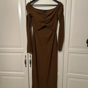 Brun långklänning från ASOS DESIGN - Snygg brun långklänning från ASOS DESIGN med lång ärm och trendig rynkad detalj framtill. Klänningen har en cut-out under bysten och är gjord i ett strukturerat, stretchigt tyg. Prislapp kvar, aldrig använd.