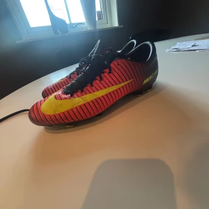 Nike Mercurial fotbollsskor med röd och gul design - Säljer ett par Nike Mercurial fotbollsskor i rött med svarta och gula detaljer. Skorna har snörning, rund tå och platt sula med dobbar för bra grepp på planen. Perfekta för dig som vill sticka ut på fotbollsplanen!