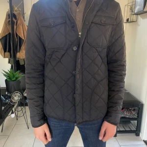 Svart quiltad jacka från Morris - Snygg svart quiltad jacka från Morris med gult innerfoder och broderad logga på ärmen. Jackan har klassisk krage, knappar framtill, två bröstfickor med lock och långa ärmar. Perfekt för dig som vill ha en stilren och tidlös look.