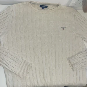 Vit kabelstickad tröja från GANT - Säljer en klassisk vit kabelstickad tröja från GANT med rund halsringning och broderad logga på bröstet. Tröjan har långa ärmar och ribbade muddar. Perfekt för en stilren och tidlös look.