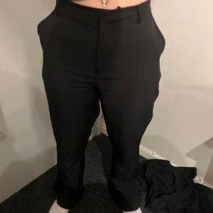 Svarta kostymbyxor med hög midja - Säljer ett par svarta kostymbyxor med hög midja och raka ben. Byxorna har fickor i sidorna och stängs med dragkedja och knapp framtill. Perfekta till en stilren look.