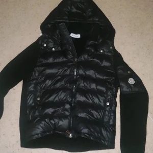 Svart pufferjacka med huva från Moncler - Säljer en svart pufferjacka från Moncler med huva och dragkedja framtill. Jackan har quiltad framsida och ärmar i stickat material. Klassisk Moncler-logga på ena ärmen och praktiska fickor med knappar.