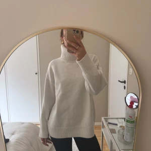 Vit polotröja i stickat material - Säljer en vit stickad polotröja med lång ärm och oversized passform. Tröjan har breda ribbade muddar vid ärmslut och nederkant samt hög polokrage. Perfekt för kyligare dagar och enkel att styla till olika outfits.