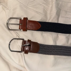 Två flätade herrbälten med brunt läder - Säljer två snygga flätade bälten med bruna läderdetaljer och silverspänne. Ett bälte är svart och det andra är grått. Perfekta accessoarer för att lyfta din outfit.det gråa är 95 cm o de blåa är 105 50 kr styck