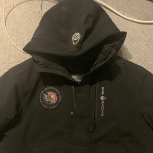 Svart Sail Racing jacka med huva - Säljer en svart jacka från Sail Racing med stor huva och dragkedja framtill. Jackan har en patch på bröstet med text och kompassmotiv samt Sail Racing-logga på bröstet. Perfekt för kyliga dagar.