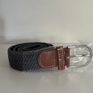 Mörkgrått flätat bälte  - Snyggt grått flätat bälte med bruna skinn-detaljer och silverspänne. Perfekt accessoar för att lyfta din outfit. Bältet har en klassisk design med kontrastsömmar på skinnpartierna.