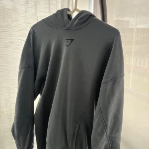 Mörkblå hoodie från Gymshark - Säljer en mörkblå hoodie från Gymshark med diskret logga framtill och stor text på ryggen. Tröjan har huva och långa ärmar, samt en bekväm passform. Perfekt för gymmet eller chill dagar. Storlek XS men sitter lika bra på S och till och med M