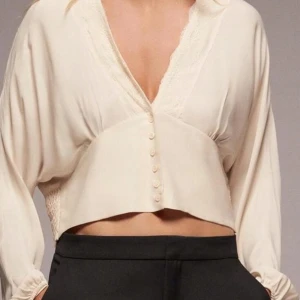 Beige långärmad topp med spets från Zara - Superfin långärmad topp från Zara i beige med v-ringning och spetsdetaljer längs halsen. Blusen har långa ärmar, knappar framtill och smockad rygg för extra komfort. Perfekt för dig som gillar romantiska och trendiga plagg.
