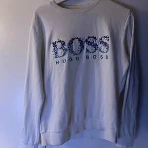 Vit sweatshirt från Hugo Boss - Vit sweatshirt från Hugo Boss med blått och turkost pixelmönstrat BOSS-tryck på bröstet. Klassisk rund halsringning och långa ärmar. Mjuk bomullskänsla och ribbade muddar vid ärmslut och nederkant. Perfekt för en clean och stilren look.