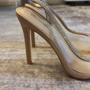 Beiga pumps med transparent rem och strassdetalj - Säljer ett par snygga pumps i beige med transparent rem och glittrande strassdetalj. Skorna har spetsig tå och hög klack, vilket ger en elegant look. Perfekta för dig som vill sticka ut lite extra. OBS!! några strass har åkt bort, inget man tänker på. 
