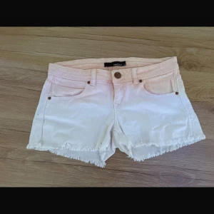Vitrosa jeansshorts med fransar - Säljer ett par vita lågmidjade jeansshorts  med ljusrosa ton upptill. Shortsen har fransade benslut, klassiska fem fickor och knappgylf. Perfekta för sommaren och har en snygg ombre-effekt.