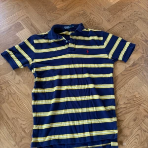 Randig pikétröja från Polo Ralph Lauren - Självklart äkta! Mörkblå pikétröja med gula och vita ränder från Polo Ralph Lauren. Klassisk krage med knappar och röd broderad logga på bröstet. Perfekt för en sportig och stilren look.(tror fläcken på sista bilden går bort i tvätten)