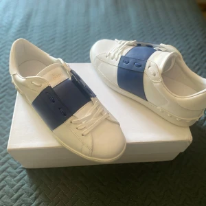 Valentino Garavani Open sneakers vit/blå - Snygga Valentino Garavani Open sneakers i vitt skinn med bred blå rem över vristen. Klassisk rund tå, vita snören och ikoniska nitar på hälen. Kommer med originalkartong och dustbag. Perfekta för dig som vill sticka ut med stil. Storlek 43. Kan diskutera pris