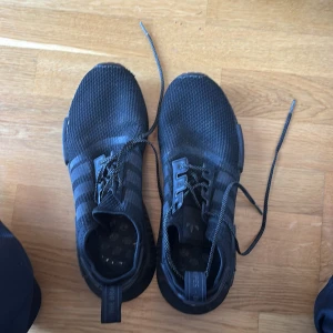 Svarta Adidas NMD sneakers - Säljer ett par svarta Adidas NMD sneakers med klassiska tre ränder på sidan. Skorna har stickad ovandel i syntetmaterial och snörning. Perfekta för dig som gillar streetstyle och vill ha bekväma skor till vardags.
