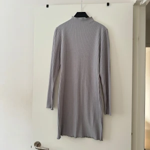 Grå ribbad klänning från Boohoo - Snygg grå ribbad kortklänning från Boohoo med lång ärm och hög hals. Klänningen har en tight passform och är tillverkad i ett mjukt, stretchigt material som sitter skönt på kroppen. 