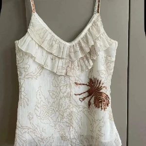 Beige blommig linnetopp med volang - Supersöt beige linnetopp med tunna axelband och volang framtill. Toppen har ett diskret blommigt mönster i beige och en cool paljettbroderad insekt på ena sidan. Perfekt för dig som gillar unika detaljer och romantisk stil.
