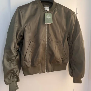 Olivgrön bomberjacka från H&M, aldrig använd, tagsen kvar. - Snygg olivgrön bomberjacka från H&M i 100% återvunnen polyester. Jackan har klassisk bomberstil med ribbade muddar, dragkedja framtill och två snedställda fickor. Helt ny. Aldrig använd.. Inköpspris 399:-, ditt pris 150:-