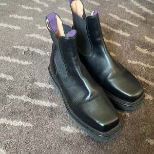 Eytys Chelsea boots - Snygga svarta chelsea boots i skinn med fyrkantig tå och elastiska paneler på sidorna. De har en cool lila flik baktill som sticker ut och gör dem unika. Perfekta för dig som vill ha en stilren men ändå personlig look.