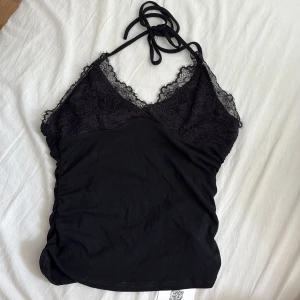 Svart spetslinnetopp från Cider - Supersnygg svart linnetopp från Cider med tunna halterneckband och v-ringad spets upptill. Öppen rygg. Toppen är figurnära och har fina spetsdetaljer över bysten som ger en extra touch. Perfekt att styla med jeans eller kjol för en trendig look.