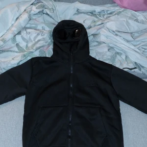 Svart hoodiejacka med goggles från C.P. Company - Svart hoodiejacka från C.P. Company med ikoniska goggles i huvan. Jackan har dragkedja framtill, två stora fickor och en clean look. Perfekt för dig som vill sticka ut med streetwear-vibe och coola detaljer.