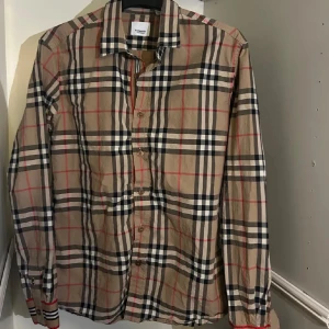 Burberry rutig skjorta i bomull - Klassisk rutig skjorta från Burberry i beige, svart, vit och röd. Skjortan har lång ärm, knappar framtill och ikoniskt rutmönster. Tillverkad i mjuk bomull och har snygga detaljer som märkta knappar och krage. Perfekt för dig som gillar stilren och tidlös design.