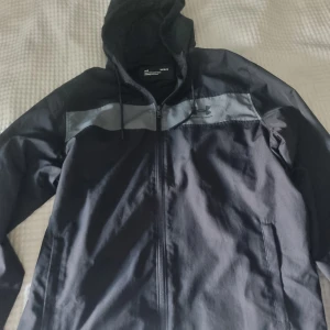 Svart jacka från Under Armour - Svart vindjacka från Under Armour med grått band över bröstet och logga. Jackan har huva med dragsko, hel dragkedja och två sidofickor. Se mått på bilder. Se gärna mina andra annonser och skapa bundels.