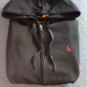 Svart hoodie från Polo Ralph Lauren - Svart hoodie från Polo Ralph Lauren med dragkedja, huva och snörning. Klassisk röd broderad logga på bröstet och känguruficka framtill. Skön passform och tillverkad i mjuk bomull, perfekt för chill dagar.