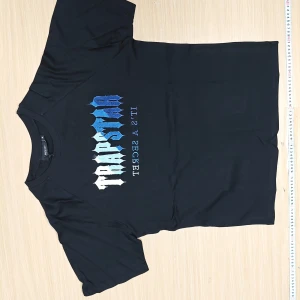 Svart Trapstar sett med blå/vit tryck - Svart t-shirt från Trapstar i storlek S med ett fett tryck i blått och vitt på bröstet. Klassisk rund hals och korta ärmar. Materialet känns mjukt och skönt, perfekt för en chill streetwear-look.